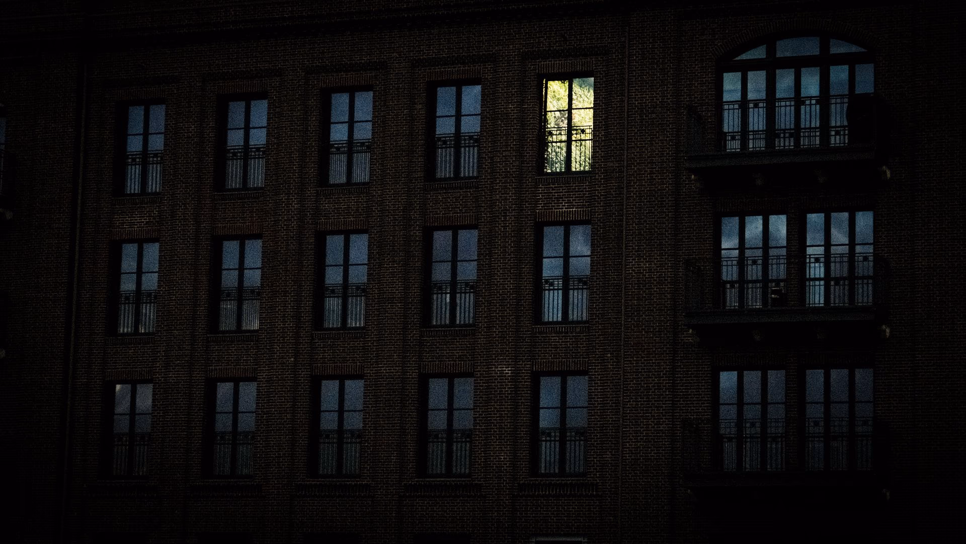 Façade sombre percée d'une seule fenêtre éclairée, lumière chaude tranchant sur le bleu nuit.