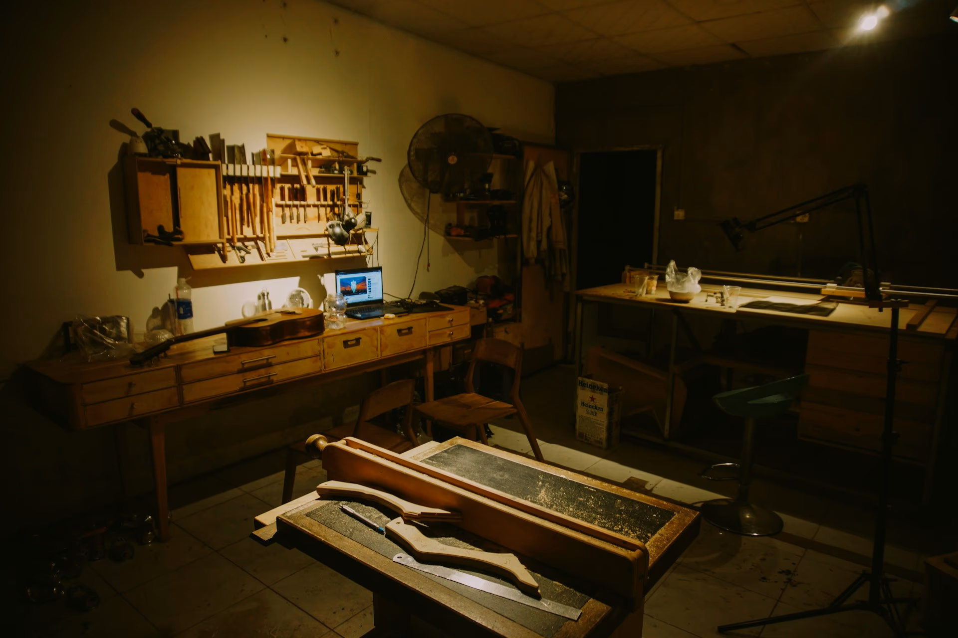 Atelier sombre rempli d'outils et de bois, établi pris dans une lumière rasante, ombres navy et reflets laiton.